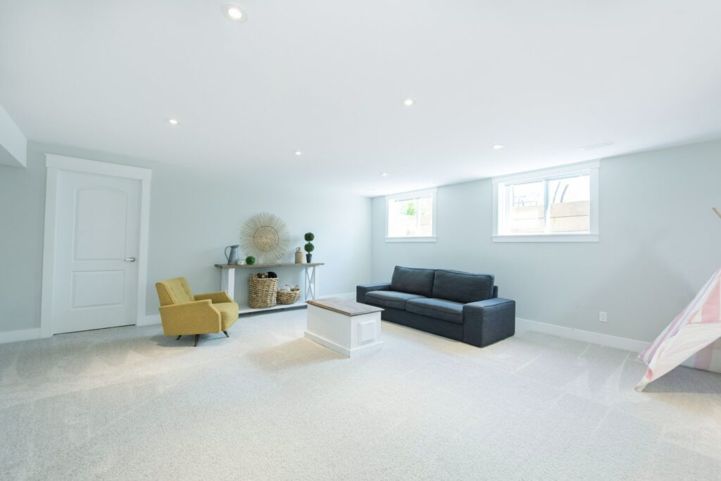 Spacious finished basement with light grey walls, large windows, modern decor, and a cozy seating area with a yellow chair and dark grey sofa.